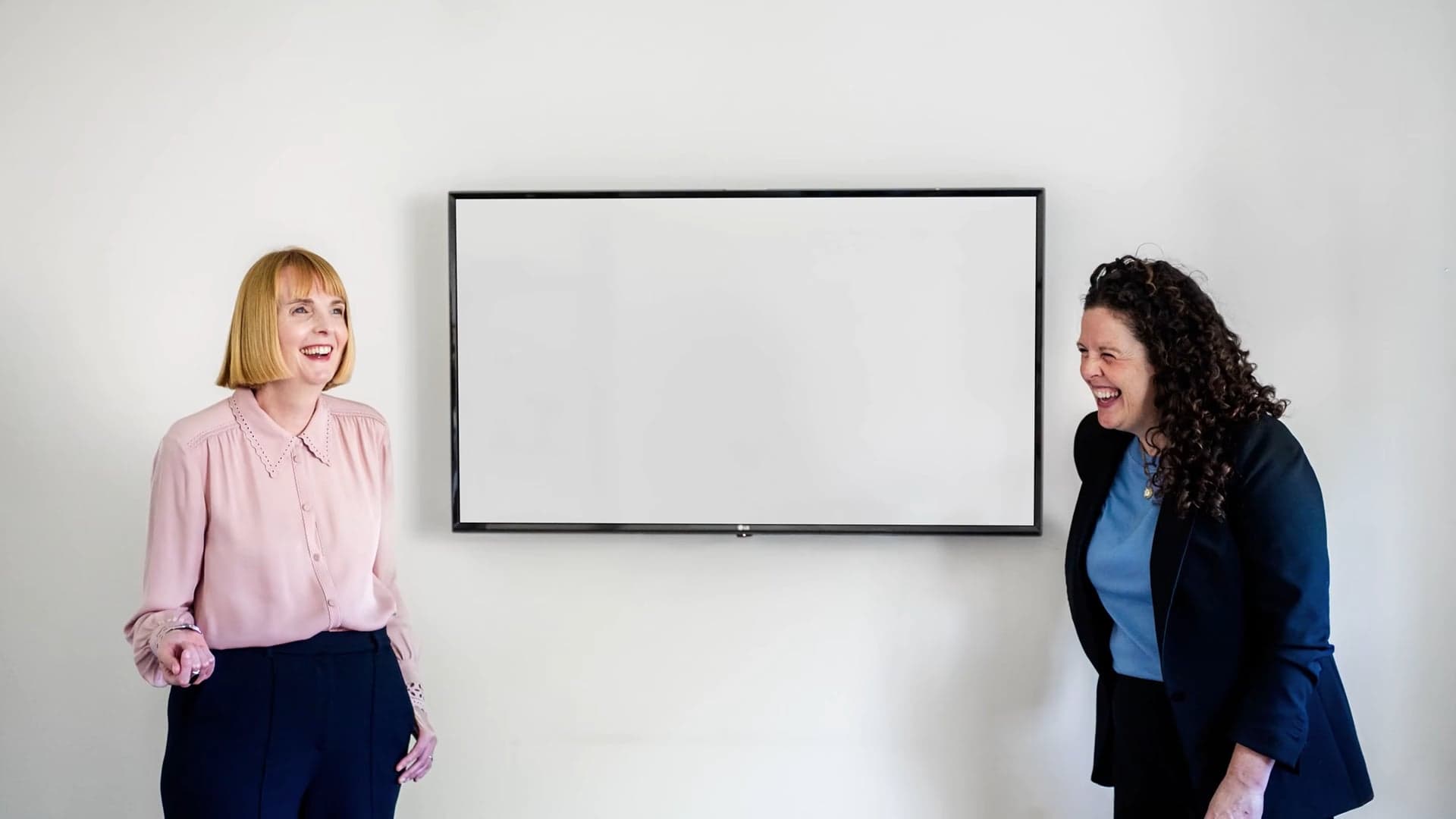 Rhi Baxter and Sara Cooke, who run We Are Brave, standing either side of a screen and laughing, from their Speaking and Event Teaser